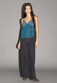 Tyrkysová saténová top s dekoratívnym šitím, skombinovaná s čiernymi širokými džínsami. Top má tenké ramienka a voľný strih.