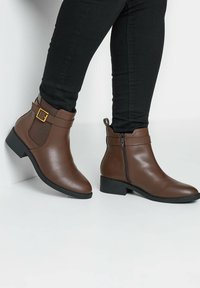 Botas tobilleras sintéticas marrones con una textura suave, que cuentan con un cierre lateral, un acento de hebilla y un tacón bajo.