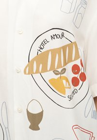 Hvid skjorte med en pladeillustration, der viser en baguette, en citronkile, cherrytomater og teksten "Hotel Amour" og "Selected."