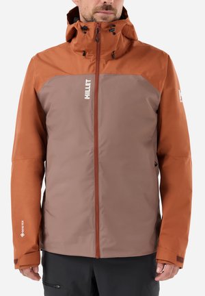 Mann trägt eine zweifarbige braune und rostfarbene wasserdichte Jacke mit Kapuze und Frontreißverschluss, steht vor einem einfarbigen hellen Hintergrund.