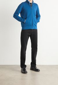 Hombre vestido con una chaqueta azul con cremallera, camisa blanca, corbata negra, pantalones vaqueros negros y zapatos de vestir negros, de pie con las manos en los bolsillos.
