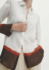 White linen shirt with a button-down front; features long sleeves with red and brown gradient accents on the cuffs and hem.