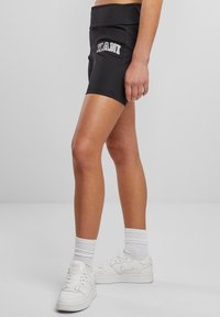 Fekete sportnadrág széles derékrésszel, rajta "KANI" logó, rugalmas anyagból készült, fehér sportcipővel és csíkos zoknival párosítva.