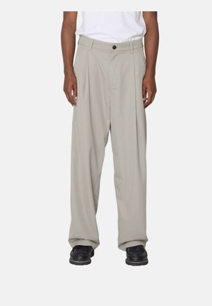 Uomo che indossa pantaloni beige larghi e scarpe nere, in piedi davanti a uno sfondo chiaro uniforme con le mani rilassate lungo i lati.