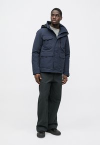 Granatowa ocieplana kurtka z kapturem, liczne kieszenie z przodu, zapięcie na zamek błyskawiczny oraz teksturowany sweter dzianinowy pod spodem. Noszona z ciemnymi spodniami i butami.