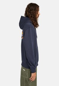 Dunkelblaues Kapuzensweatshirt aus Baumwollmix; mit Fronttasche, gerippten Bündchen, Saum und buntem grafischen Design auf dem Rücken.