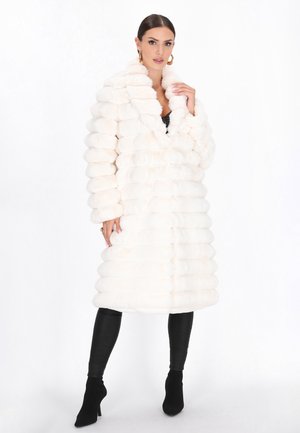 Cappotto in pelliccia sintetica bianca con un design strutturato, righe orizzontali, un ampio colletto e una lunghezza media. Indossato sopra pantaloni neri aderenti.
