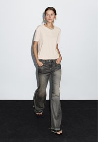 Beige kortærmet skjorte og mørkegrå flared jeans. Jeansene har frynset kant, slidt stofstruktur, slidt farvegradient og knaplommer.