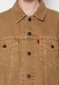 Chaqueta de mezclilla color crema con cuello camisero, botones negros y dos bolsillos en el pecho. Presenta costuras visibles y una pequeña etiqueta de la marca.