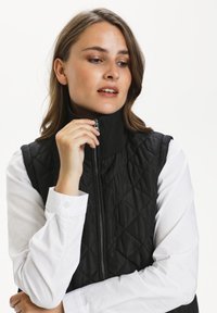 Gilet matelassé noir avec un col haut et une fermeture éclair, porté sur une chemise blanche à boutons avec des détails aux poignets. Texture lisse.