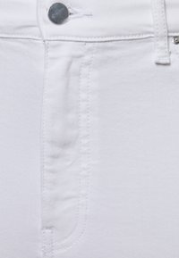 Tejido de mezclilla blanca mostrando un primer plano del costura, un botón de metal y el contorno de un bolsillo. La textura parece suave con un ligero estiramiento.