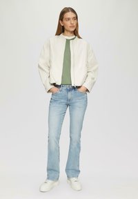 Lekka, kremowa bomberka z kołnierzem noszona na zielonej bluzce, w połączeniu z jasnoniebieskimi spodniami w prostym kroju i białymi sneakersami.