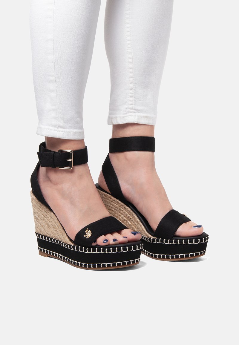 Černé klínové sandály s plátěným svrškem, tkanou juty platformou a dekorativním bílým prošíváním. Nastavitelný náramek na kotník a malý zlatý akcent.