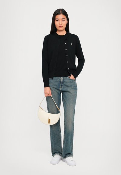Donna che indossa un cardigan nero e jeans, con una borsa bianca in mano, in piedi contro uno sfondo bianco uniforme.