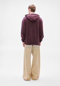 Felpa oversize color vinoso con cappuccio a coulisse, abbinata a pantaloni beige larghi con tasche posteriori e dettaglio in denim.