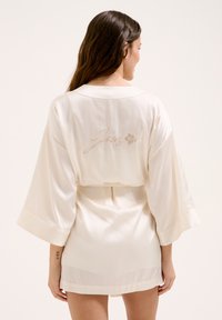 Raso crema con una vestibilità comoda, maniche ampie e cintura in vita. Presenta ricamato "Juicy" con accenti floreali sul retro.