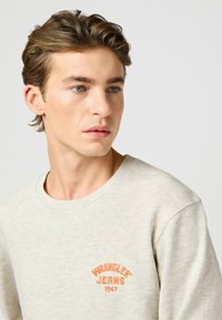 Felpa in cotone grigio chiaro con girocollo a costine e logo ricamato arancione "WRANGLER JEANS 1947" sul lato sinistro del petto.