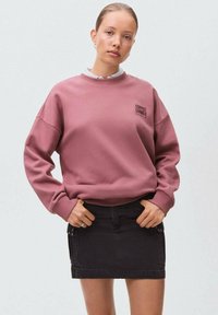 Rosa Sweatshirt mit gerippten Bündchen und Kragen, mit einem kleinen aufgedruckten Logo. Getragen mit einem schwarzen Denimrock, knöchellang.