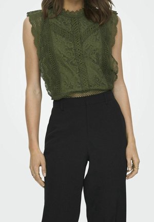 Femme portant un haut en dentelle verte sans manches avec des bords festonnés et un pantalon noir taille haute, debout, les bras détendus le long du corps.
