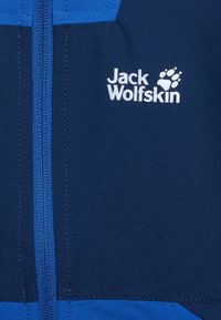Detailní záběr na přední stranu modré bundy s vertikálním zipem a bílým logem Jack Wolfskin, které obsahuje text a otisk tlapky.
