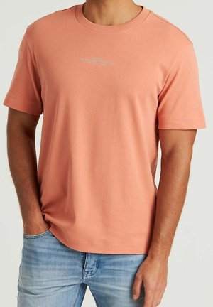 Mężczyzna ubrany w łososiowy t-shirt z krótkim rękawem z małym białym napisem na piersi, w połączeniu z jasnoniebieskimi dżinsami, ręka w kieszeni.