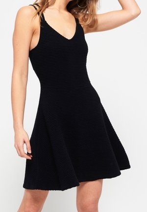 Kvinde iført en ærmeløs sort tekstureret A-formet kjole med tynde stropper og V-udskæring, poserer med den ene arm løftet.