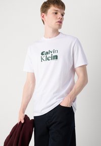 Hvit bomull t-skjorte med et sentralt grønt "Calvin Klein"-logo. Korte ermer, rund hals, avslappet passform, kombinert med mørke bukser og en burgunder genser.