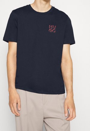 Personne portant un T-shirt noir à manches courtes avec un petit logo rouge "HUGO" sur la poitrine et un pantalon beige, debout devant un fond uni.