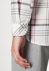 Main reposant sur le côté portant une chemise à carreaux blanche avec des lignes roses et noires et un pantalon gris clair texturé.