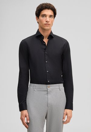 Mann mit lockigem dunklem Haar, der ein schwarzes, geknöpftes Hemd trägt, das in hellgraue Anzughosen gesteckt ist, stehend vor einem einfachen Hintergrund.