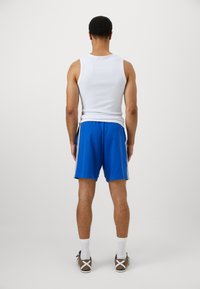 Λευκό αμάνικο μπλουζάκι με μπλε σορτσάκια που έχουν πλευρικές λωρίδες, εμφανίζεται από πίσω. Καφέ αθλητικά παπούτσια με λευκό σχέδιο σταυρού και κάλτσες μέχρι τον αστράγαλο.