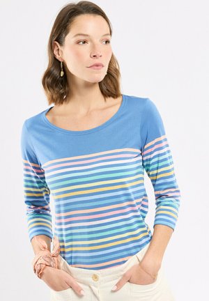 Femme aux cheveux bruns courts portant un chemisier bleu à manches longues avec des rayures horizontales multicolores, un pantalon blanc et un foulard à motifs pêche autour du poignet.