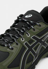 Chaussure de sport noire et vert olive avec un matériau en maille, des accents texturés, des lacets noirs avec des détails blancs et un logo réfléchissant.