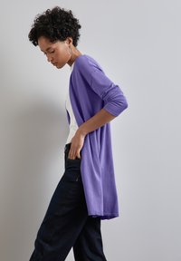 Lång, öppen cardigan i mjuk lila stickning, kombinerad med en vit topp och mörka jeans. Har uppvikta ärmar och en avslappnad silhuett.