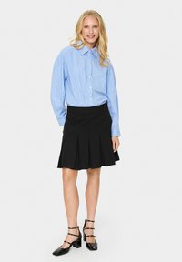 Chemise boutonnée rayée bleu clair avec un col, associée à une jupe plissée noire taille haute et des chaussures à talons carrés noires.