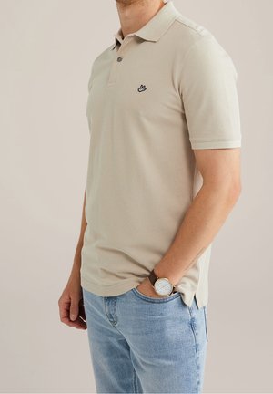 Beżowa koszulka polo z teksturowanym splotem, krótkie rękawy i czarnym logo na piersi. W połączeniu z jasnoniebieskimi dżinsami i brązowym zegarkiem.