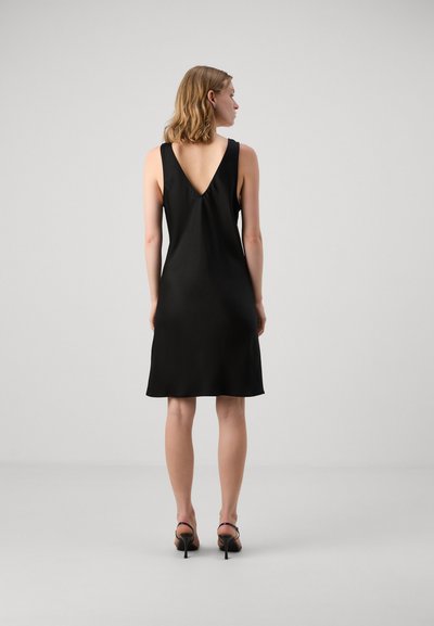 Robe noire sans manches avec un dos en V, en tissu lisse et à coupe décontractée, se terminant juste au-dessus du genou. Portée avec des sandales à talons noires.