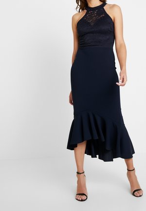 Kvinde iført en ærmeløs navyblå kjole med blonder på overdelen, høj halsudskæring og asymmetrisk flæsekanter, kombineret med sorte ankelrem-sandaler.