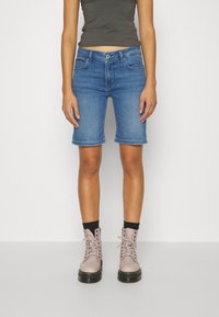 Persona vistiendo pantalones cortos de mezclilla azules de longitud media, calcetas negras, botas claras de encaje rosa y una camiseta sin mangas gris oscuro contra un fondo blanco.