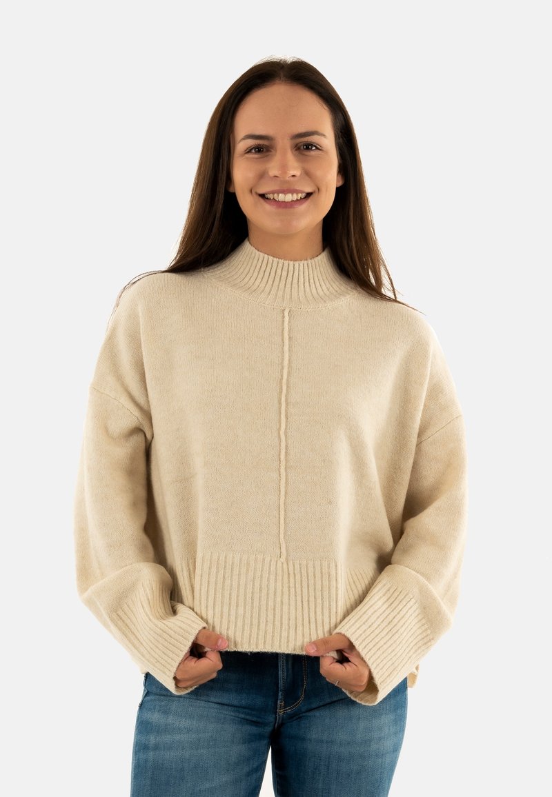 Pull en tricot texturé couleur crème, avec un col haut à côtes et un ourlet large à côtes. Présente des épaules tombantes et de longues manches.