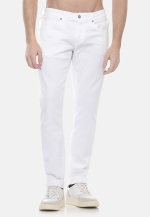 Mann trägt schlanke weiße Jeans und weiße Turnschuhe, steht vor einem hellen, schlichten Hintergrund mit entspannt an den Seiten hängenden Armen.