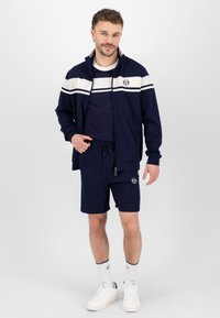 Marineblå idrettsdrakt med jakke med glidelås, som har hvite striper og logo, matchende shorts og hvite joggesko. Glatt tekstur, uformell design.