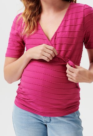 Haut fuchsia en tissu texturé avec manches courtes et décolleté en V profond, doté d'un détail noué à la taille. Associé à un jean bleu.