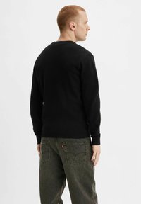 Maglione nero con scollatura a girocollo e polsini a costine, abbinato a jeans verde scuro larghi con tasche posteriori.