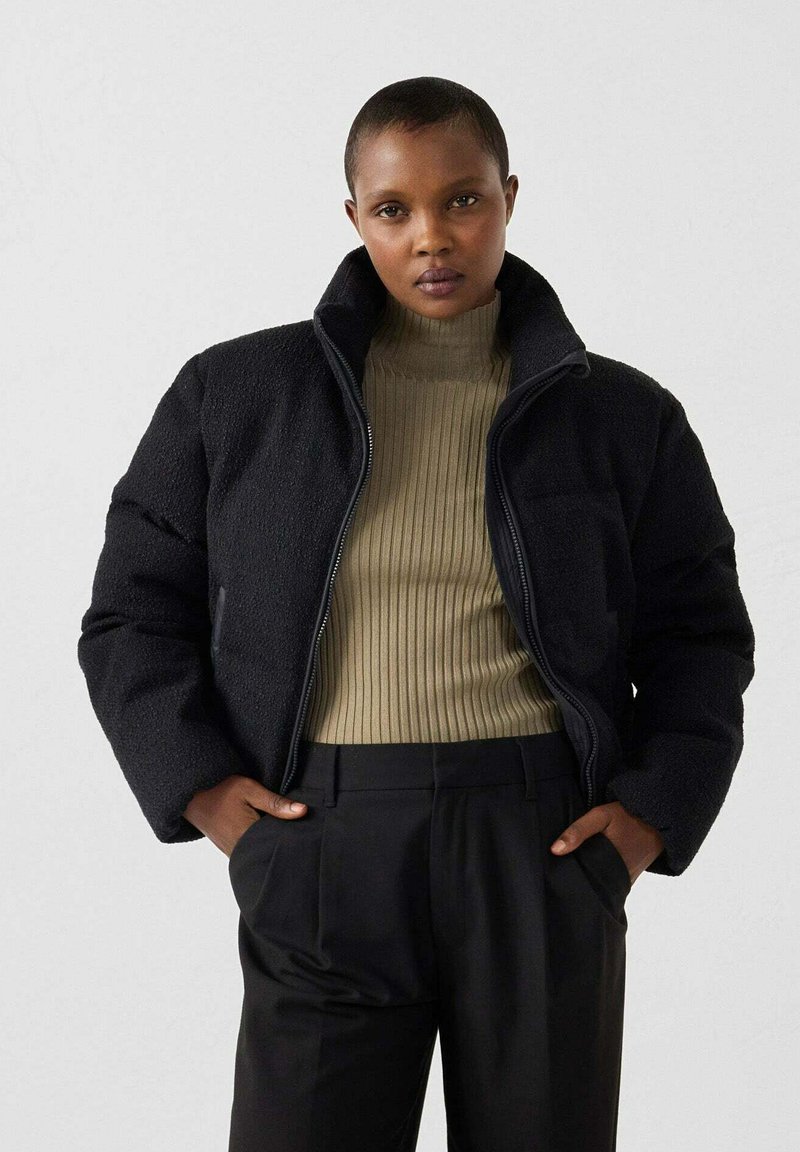 Personne portant une doudoune noire par-dessus un pull col roulé beige à côtes et un pantalon noir, se tenant les mains dans les poches.