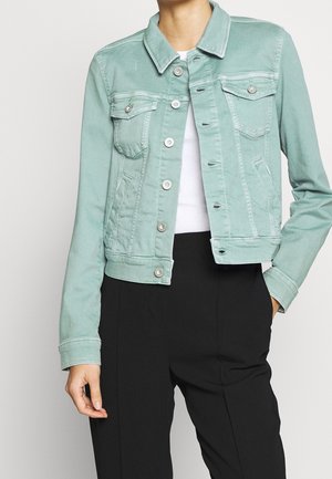 Frau trägt eine hellgrüne Jeansjacke über einem weißen Top und schwarzen Hosen, mit einer Hand in der Tasche und der Jacke teilweise zugeknöpft.