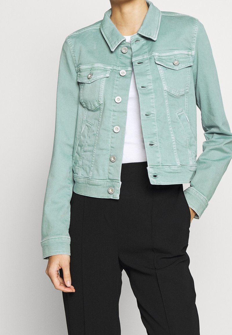 Kvinde iført lysegrøn denimjakke over hvid top og sorte bukser, med den ene hånd i lommen og jakken delvist knappet.