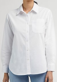 Chemise blanche boutonnée avec col, manches longues, une poche poitrine, et un ourlet droit. Fabriquée dans un tissu texturé avec une finition lisse.