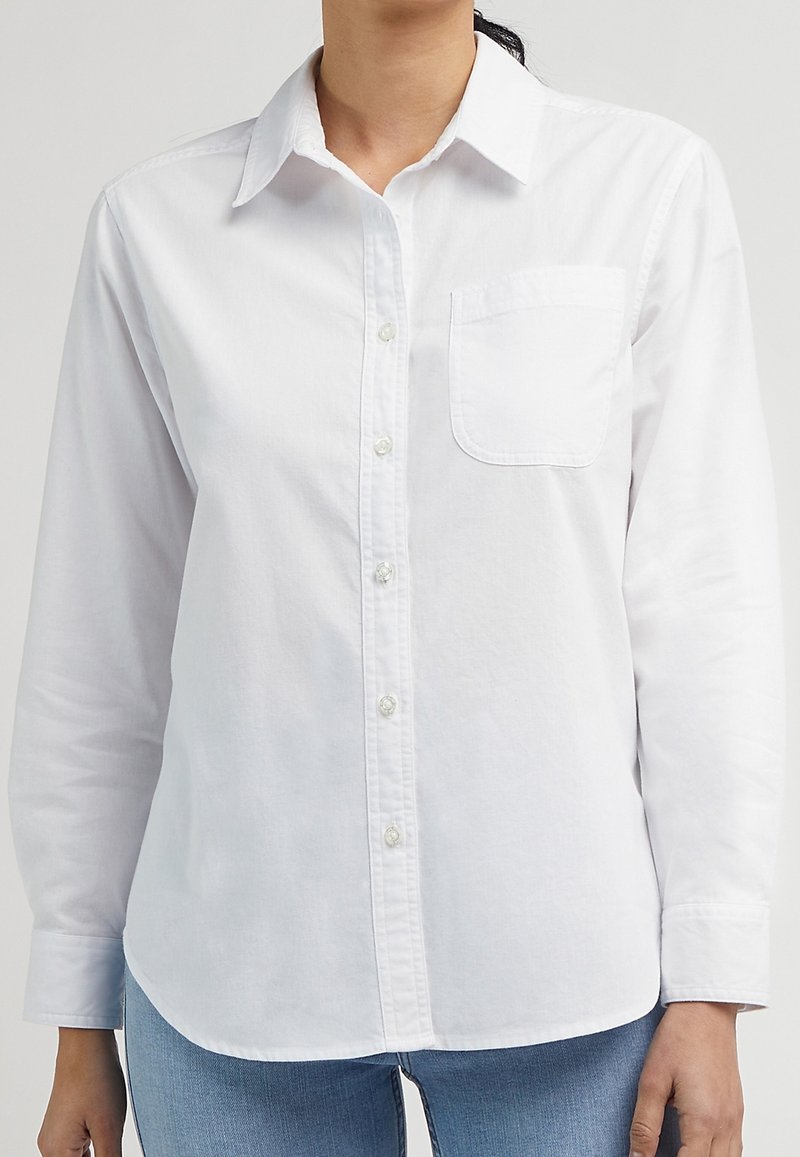 Chemise blanche boutonnée avec col, manches longues, une poche poitrine, et un ourlet droit. Fabriquée dans un tissu texturé avec une finition lisse.
