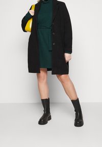 Cappotto nero con grandi bottoni, indossato sopra un vestito verde scuro con una cintura in vita. Abbinato a stivali neri fino al ginocchio e a una borsa gialla.